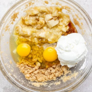 A glass bowl containing mashed bananas, two eggs, yogurt, brown sugar, and a liquid ingredient.