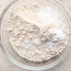 A clear glass bowl containing a mixture of flour and baking powder on a textured beige surface.