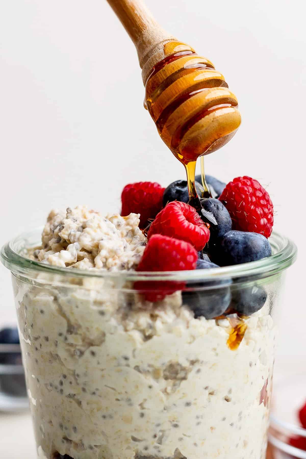 A glass jar of cheesecake overnight oats topped with blueberries and raspberries, drizzled with honey from a wooden dipper.