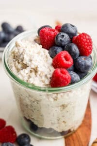 A jar of cheesecake overnight oats topped with fresh blueberries and raspberries sits on a wooden surface, offering a tempting twist. More berries are visible in the background, adding bursts of color and flavor.