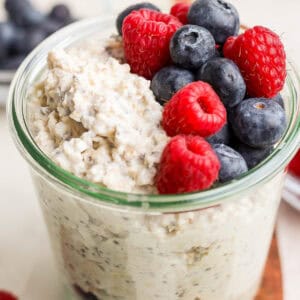 A jar of cheesecake overnight oats topped with fresh blueberries and raspberries sits on a wooden surface, offering a tempting twist. More berries are visible in the background, adding bursts of color and flavor.