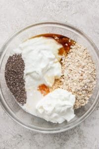 A clear bowl containing cheesecake overnight oats, yogurt, chia seeds, honey, and milk rests on a textured surface.