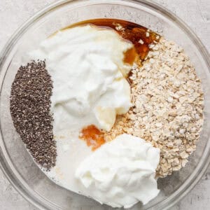 A clear bowl containing cheesecake overnight oats, yogurt, chia seeds, honey, and milk rests on a textured surface.