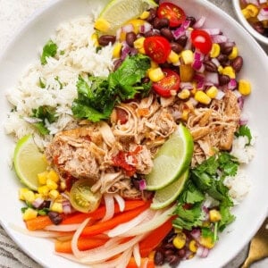 A bowl with shredded chicken, rice, black beans, corn, red onion, tomatoes, lime wedges, cilantro, and sliced bell peppers.