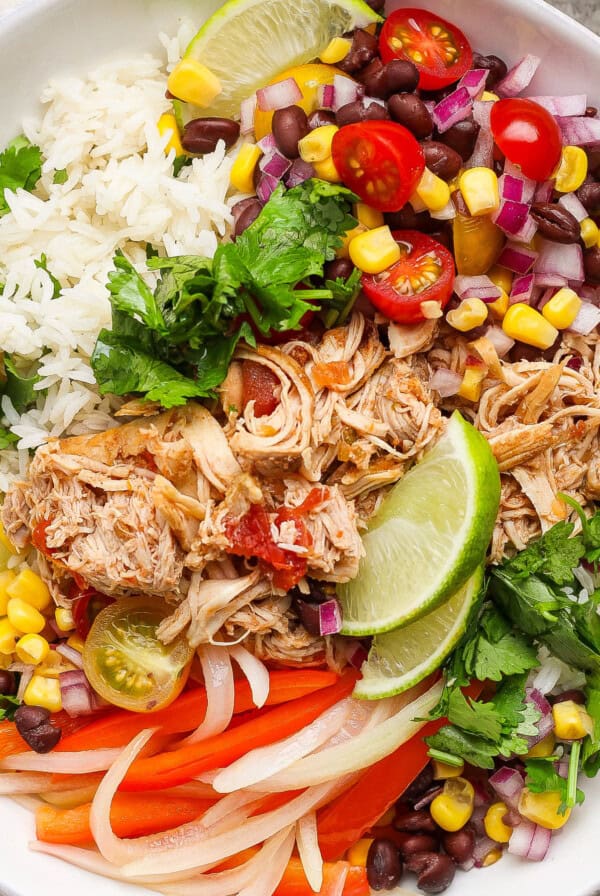 A bowl with shredded chicken, rice, black beans, corn, red onion, tomatoes, lime wedges, cilantro, and sliced bell peppers.