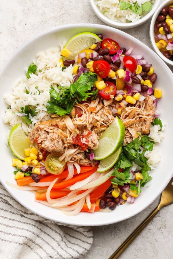 A bowl of rice topped with shredded chicken, black beans, corn, tomatoes, onions, lime wedges, and cilantro, garnished with sliced peppers.