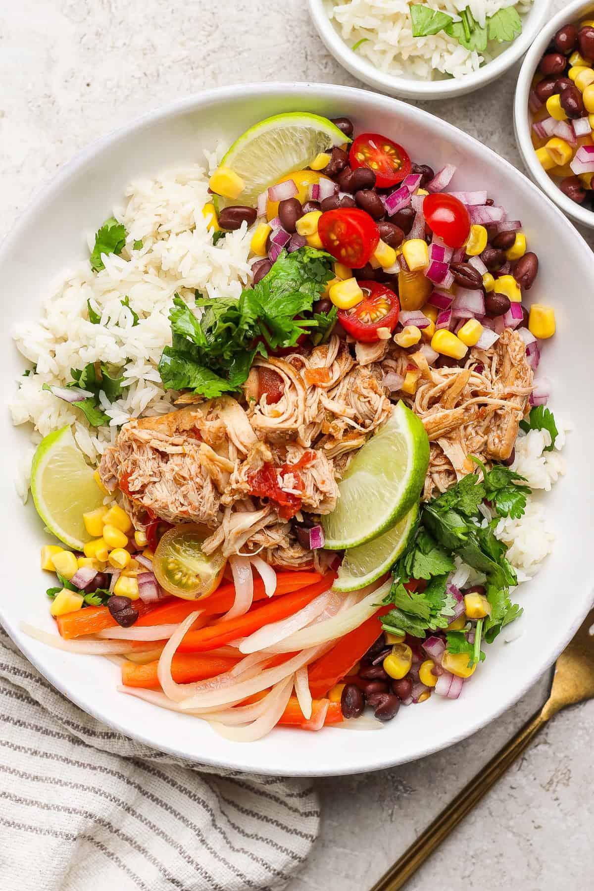 A bowl of rice topped with shredded chicken, black beans, corn, tomatoes, onions, lime wedges, and cilantro, garnished with sliced peppers.