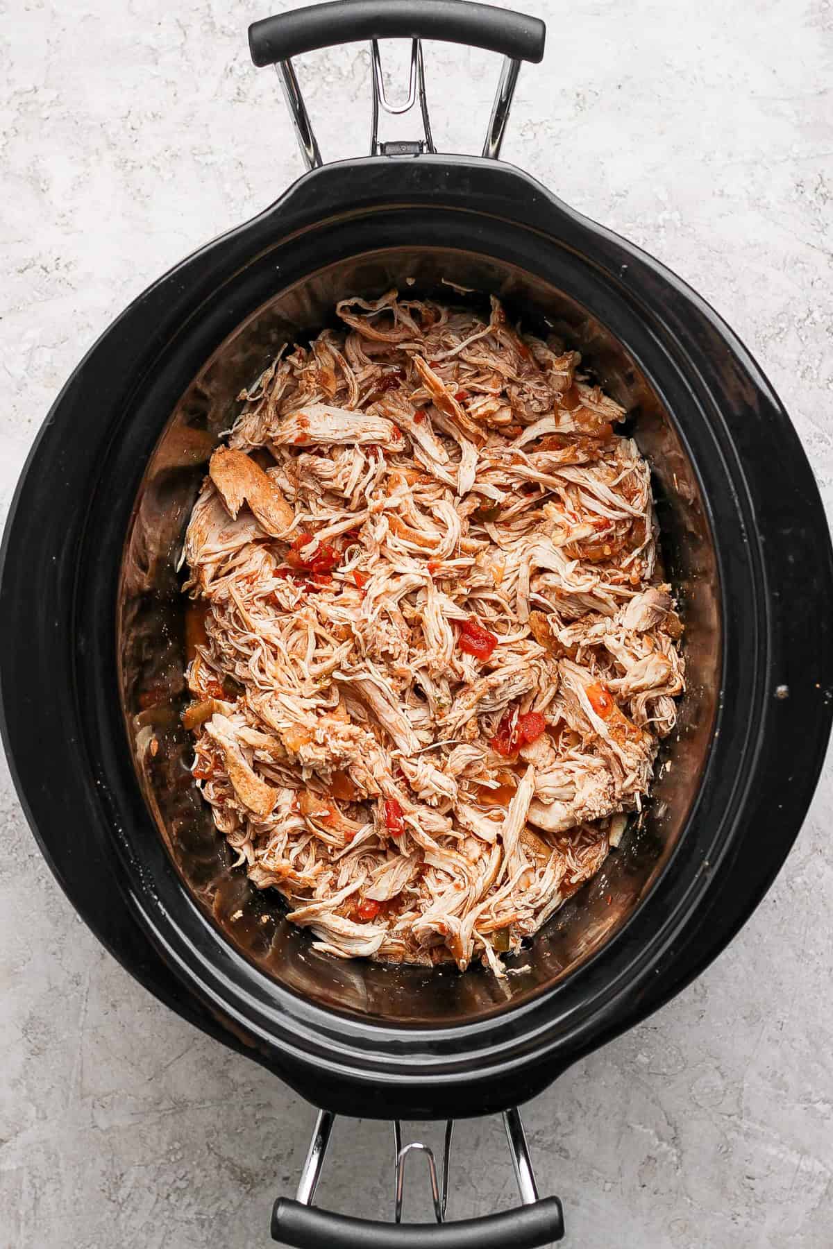 Shredded chicken mixed with red peppers in a slow cooker on a gray textured surface.