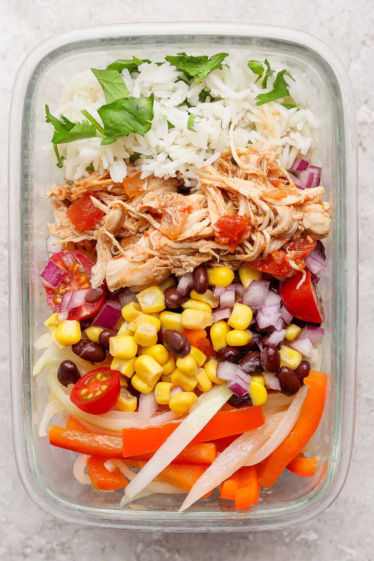 A glass container with rice, shredded chicken, black beans, corn, red onions, cherry tomatoes, sliced bell peppers, and cilantro.