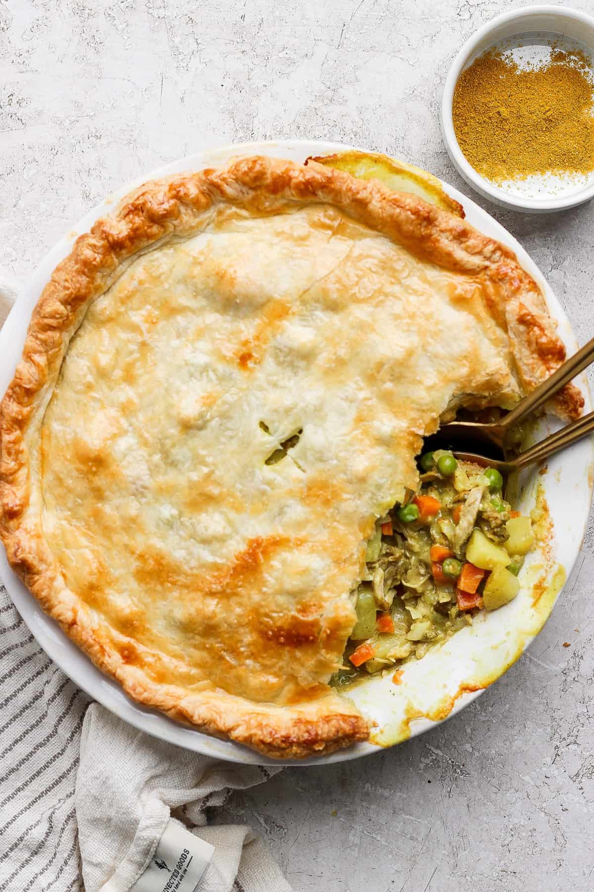 A savory pie with a golden crust is partially sliced, revealing a filling of vegetables and chicken. Two spoons are in the pie, and a bowl of seasoning is nearby.