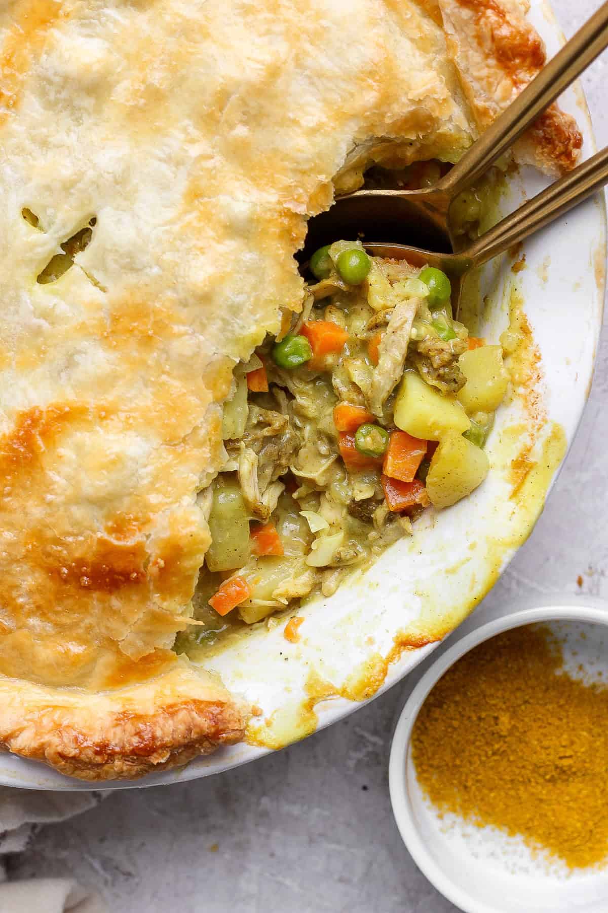 A pot pie with a flaky crust partially open, revealing a filling of chicken, carrots, peas, and potatoes. Two spoons are placed inside, next to a small dish of curry powder.