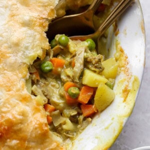 A close-up of a pot pie with a golden crust, showing a filling of chicken, peas, carrots, and potatoes, with two spoons partially inside.