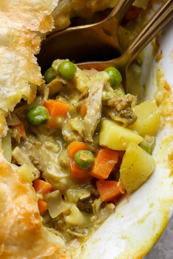 A close-up of a pot pie with a golden crust, showing a filling of chicken, peas, carrots, and potatoes, with two spoons partially inside.