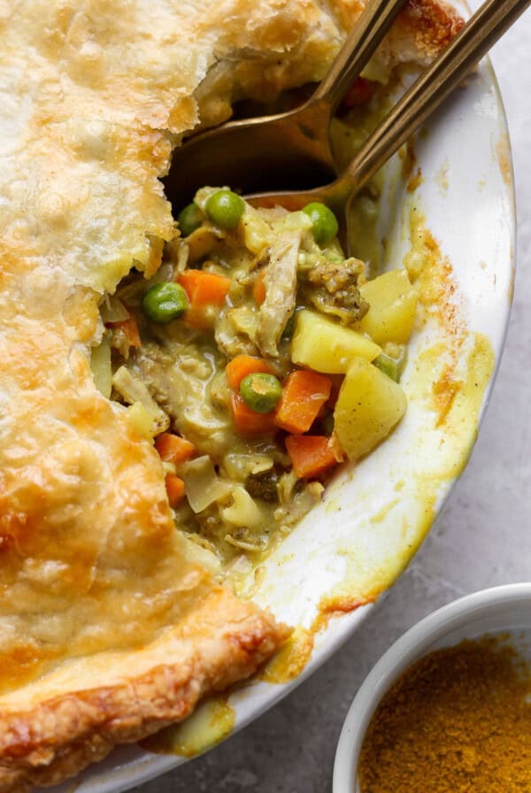 Close-up of a pot pie with flaky golden crust, filled with chicken, peas, carrots, and potatoes. A spoon is placed in the pie, and a small bowl of seasoning is nearby.