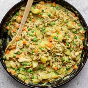 A skillet of cooked chicken, potatoes, carrots, and peas mixed in a creamy sauce, with a wooden spoon on the side.