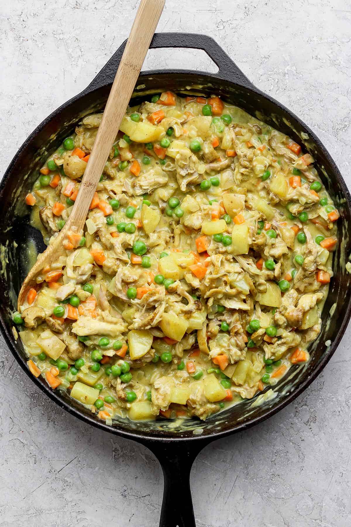 A skillet of cooked chicken, potatoes, carrots, and peas mixed in a creamy sauce, with a wooden spoon on the side.