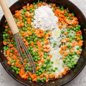 A skillet with sautéed carrots, peas, onions, butter, and a pile of flour, stirred with a whisk.