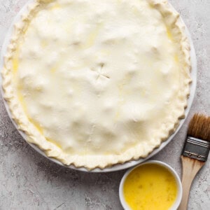 Unbaked pie with a crimped edge on a round dish, accompanied by a small bowl of egg wash and a pastry brush on a textured surface.