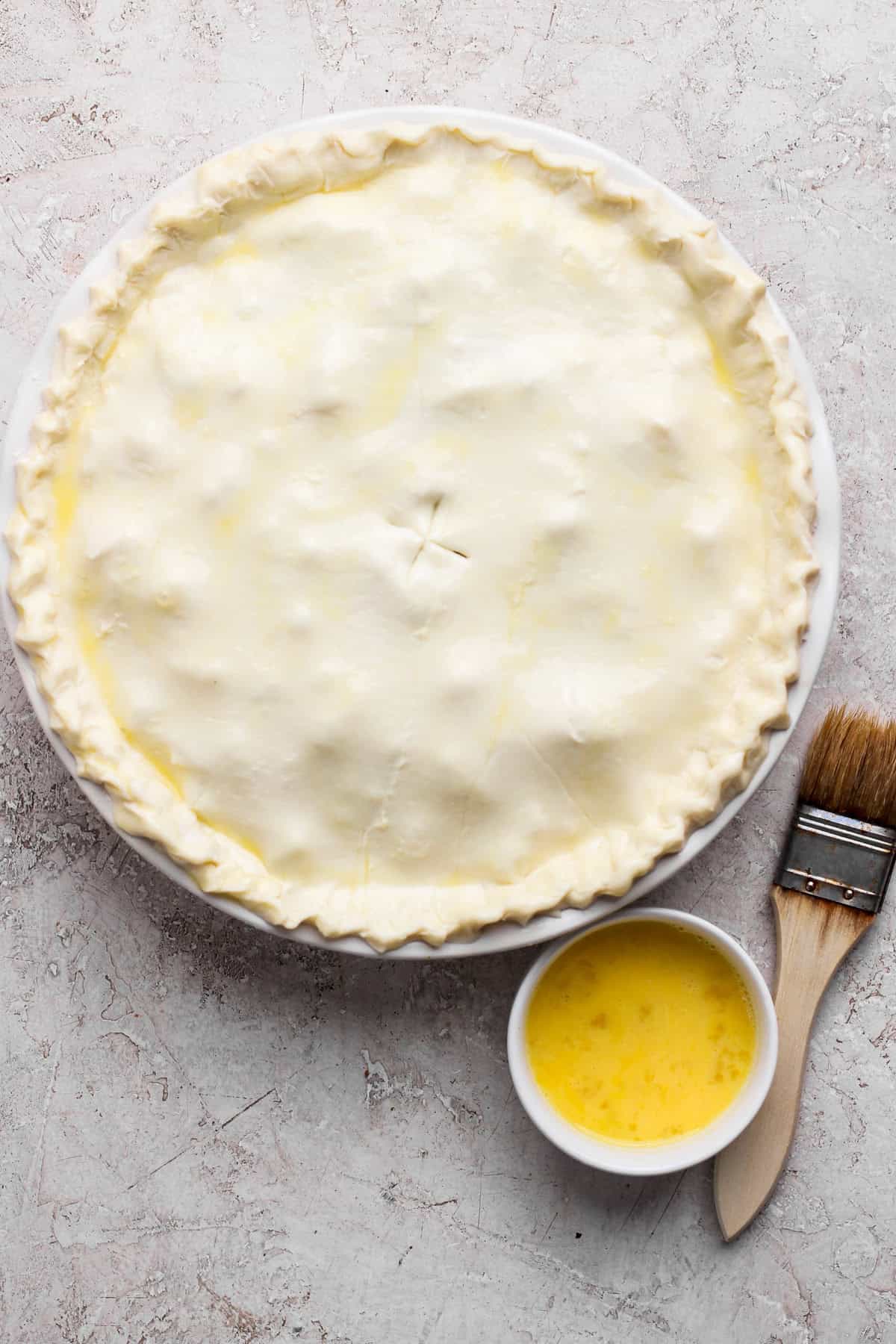 Unbaked pie with a crimped edge on a round dish, accompanied by a small bowl of egg wash and a pastry brush on a textured surface.