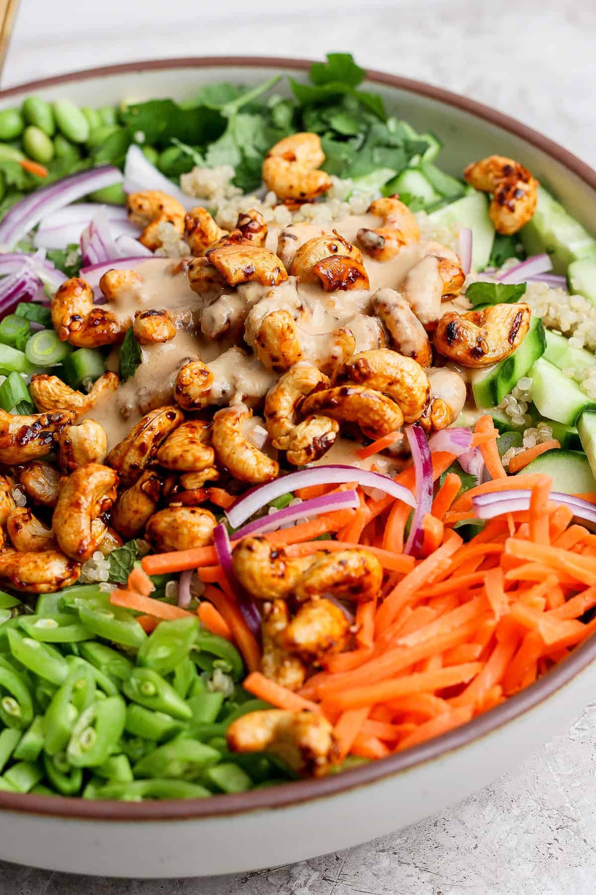 A colorful salad with cashews, quinoa, cucumbers, red onions, sliced carrots, green beans, and greens, topped with a creamy dressing, served in a bowl.