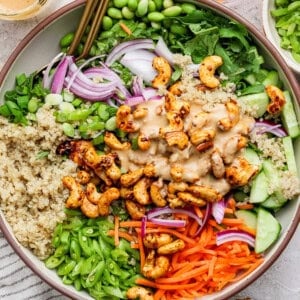 A bowl of salad with quinoa, edamame, onions, cucumbers, carrots, cashews, and green onions, topped with a creamy dressing.