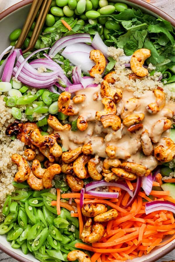 A bowl of salad with quinoa, edamame, onions, cucumbers, carrots, cashews, and green onions, topped with a creamy dressing.