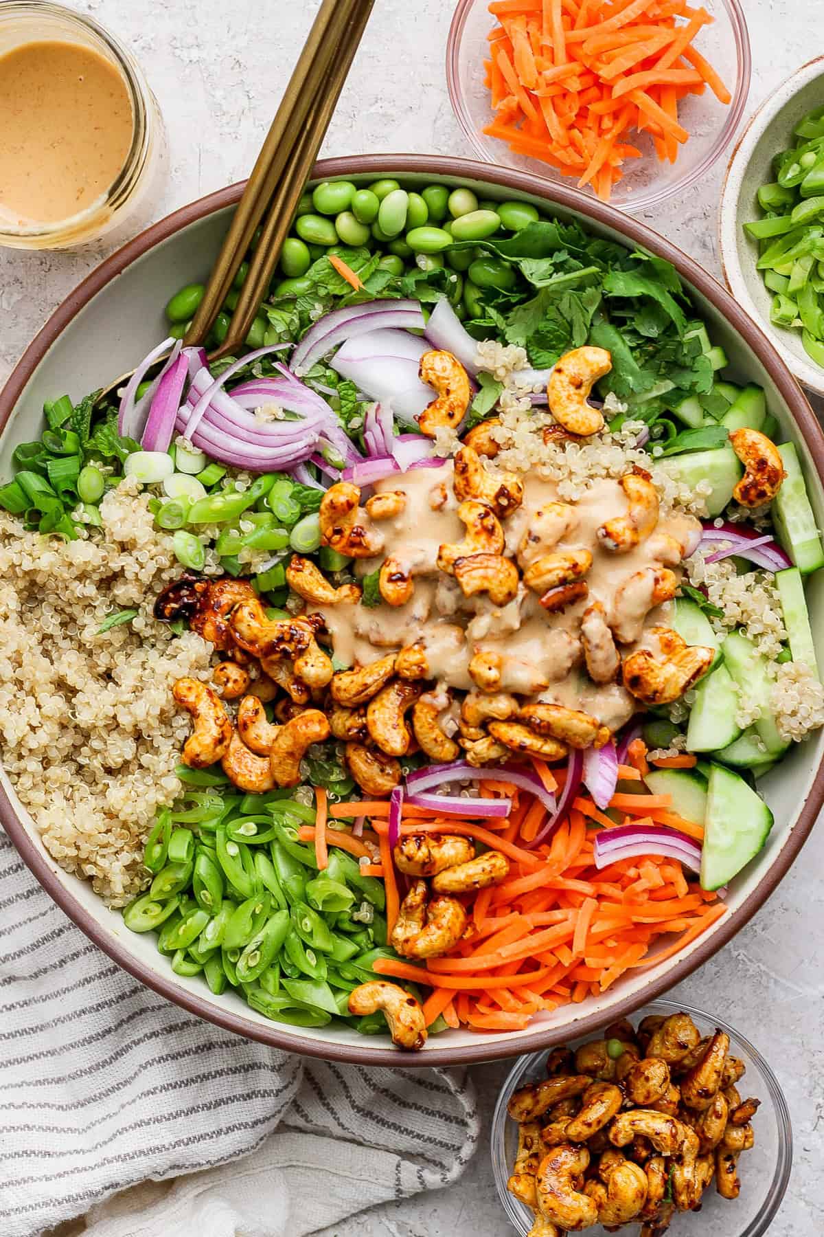 A bowl containing quinoa, cashews, sliced red onions, carrots, cucumbers, edamame, and greens, topped with a creamy dressing. Additional ingredients are in small bowls around it.