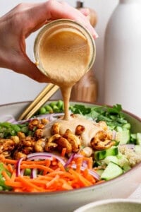 A hand pours creamy dressing over a salad with carrots, cucumbers, red onions, roasted chickpeas, and greens in a bowl.