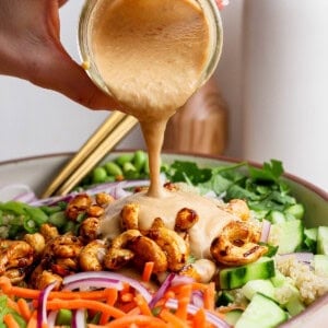 A hand pours creamy dressing over a salad with carrots, cucumbers, red onions, roasted chickpeas, and greens in a bowl.