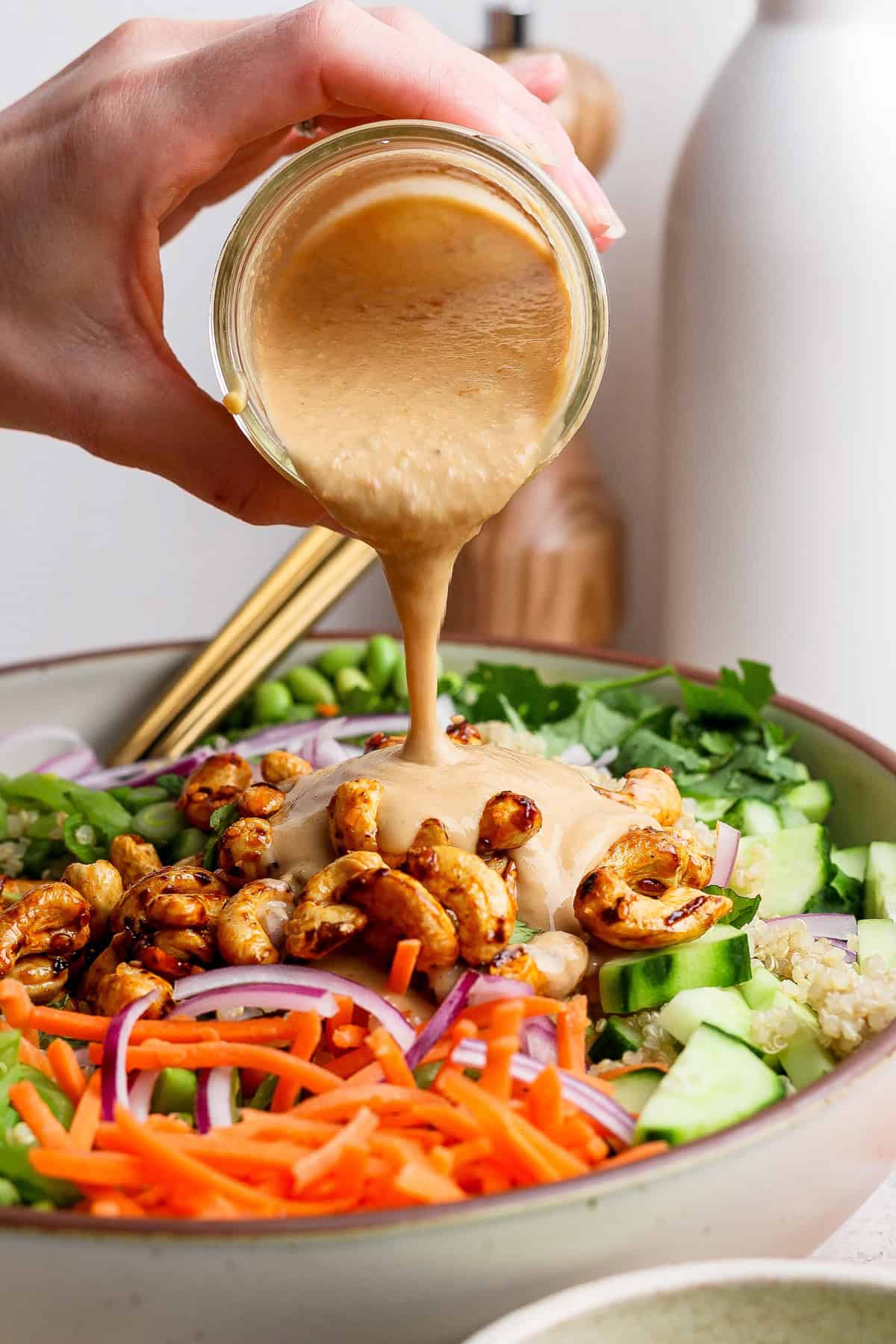 A hand pours creamy dressing over a salad with carrots, cucumbers, red onions, roasted chickpeas, and greens in a bowl.