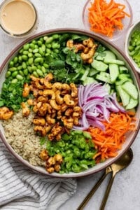 A colorful salad in a bowl includes cashews, edamame, quinoa, sliced cucumbers, shredded carrots, red onions, and herbs. A small bowl of dressing and chopped carrots are nearby.