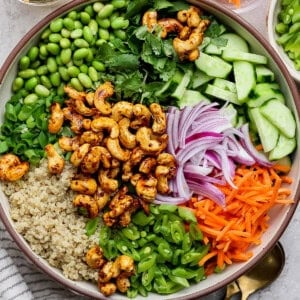 A colorful salad in a bowl includes cashews, edamame, quinoa, sliced cucumbers, shredded carrots, red onions, and herbs. A small bowl of dressing and chopped carrots are nearby.