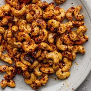 A plate of roasted cashews seasoned with chili flakes and salt on a light gray surface.