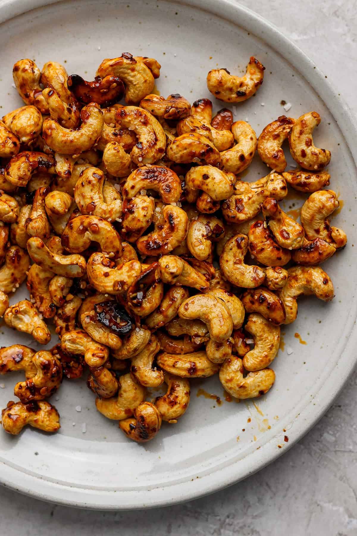 A plate of roasted cashews seasoned with chili flakes and salt on a light gray surface.