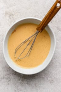 A whisk in a white bowl containing a pale orange mixture on a speckled surface.