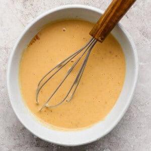 A whisk in a white bowl containing a pale orange mixture on a speckled surface.