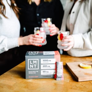 Three friends enjoying drinks, including a refreshing sleepy girl mocktail, sit beside a box of LMNT electrolyte drink mix on the wooden table.