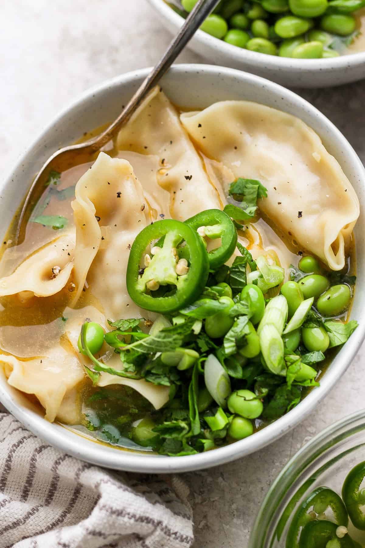 A bowl of soup with dumplings, sliced jalapeños, green onions, edamame, and cilantro. A spoon rests in the bowl, and a striped cloth is nearby.