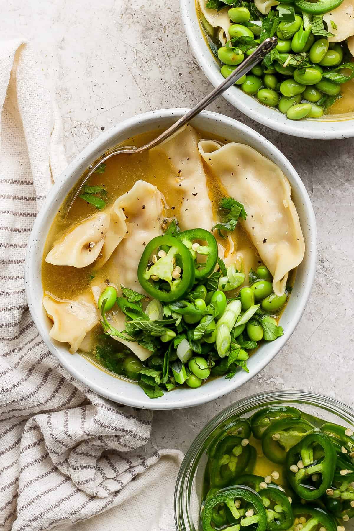 A bowl of dumpling soup garnished with sliced jalapeños, chopped green onions, and cilantro, with a side of edamame. A striped cloth is beside the bowl.