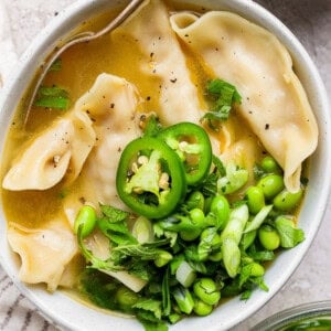 A bowl of soup with dumplings, topped with sliced jalapeños, edamame, green onions, and cilantro.