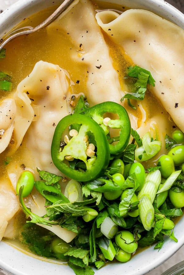 A bowl of soup with dumplings, topped with sliced jalapeños, edamame, green onions, and cilantro.