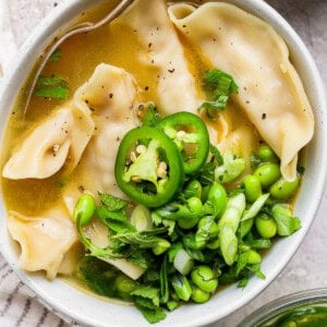 A bowl of broth with dumplings, sliced green chili, edamame, and chopped green onions, garnished with herbs. A spoon is resting in the bowl.