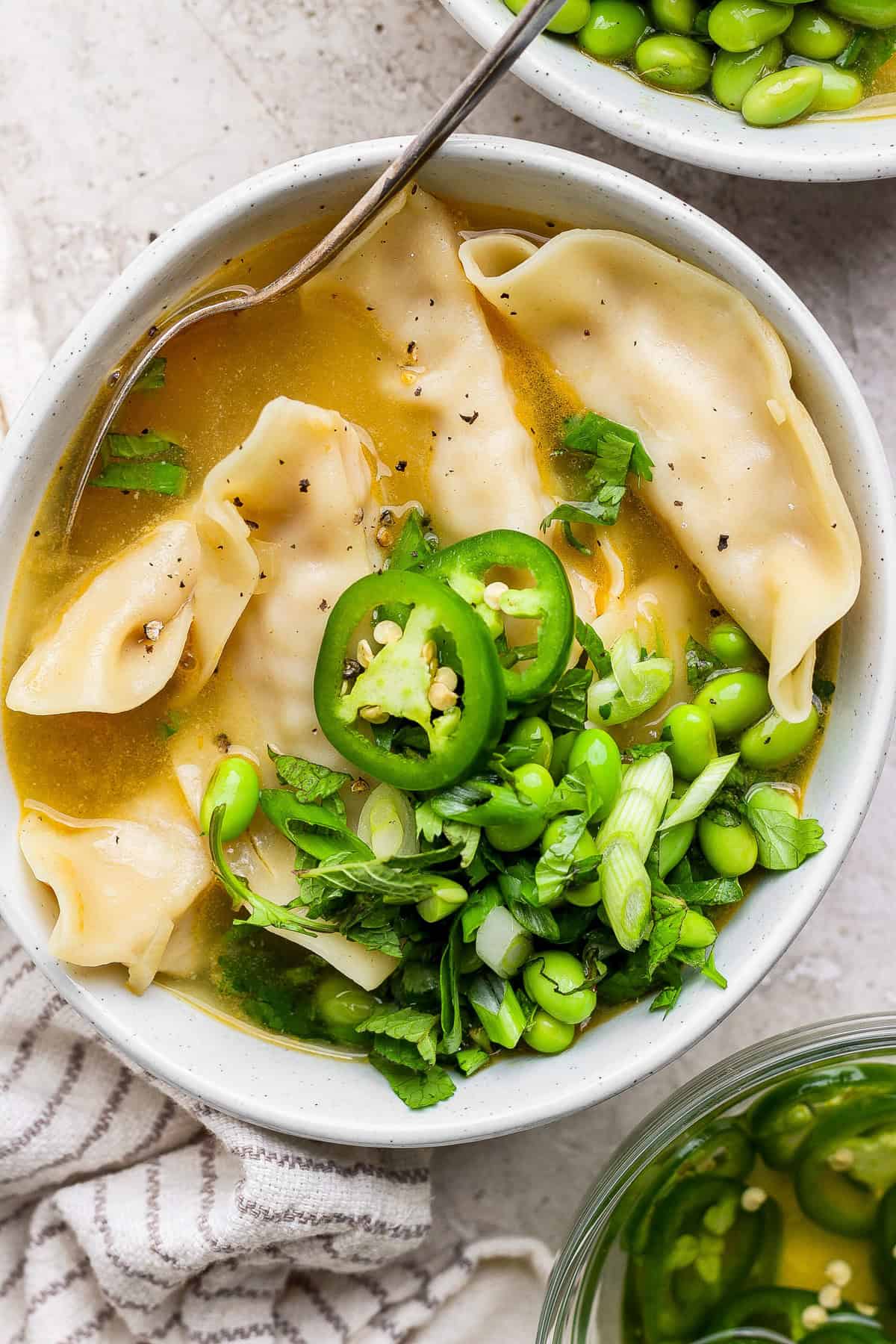 A bowl of broth with dumplings, sliced green chili, edamame, and chopped green onions, garnished with herbs. A spoon is resting in the bowl.