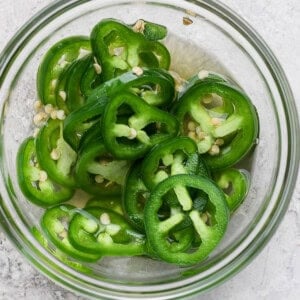 A glass bowl filled with sliced green jalapeños on a light gray surface.