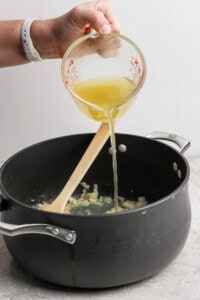 A person pours yellow liquid from a measuring cup into a black pot containing sautéed onions, with a wooden spoon resting in the pot.
