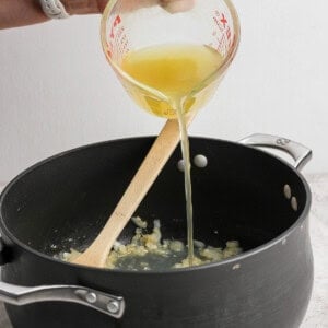 A person pours yellow liquid from a measuring cup into a black pot containing sautéed onions, with a wooden spoon resting in the pot.