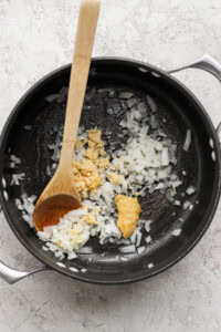 Chopped onions and minced garlic sauté in a black pot with a wooden spoon on a gray textured surface.