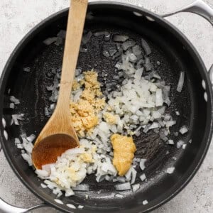 Chopped onions and minced garlic sauté in a black pot with a wooden spoon on a gray textured surface.