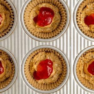 Muffin tin filled with unbaked muffin batter topped with red jam, placed in paper liners, ready for baking.