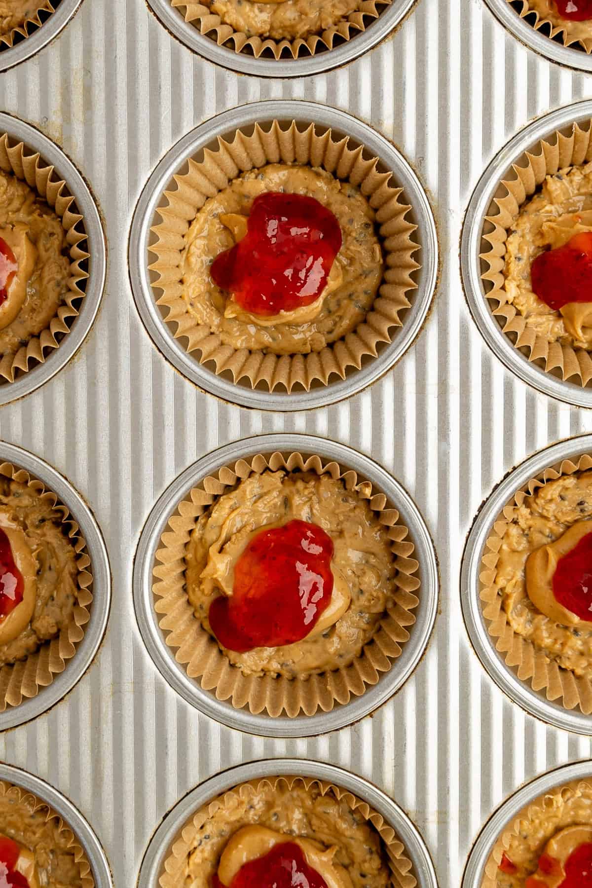 Muffin tin filled with unbaked muffin batter topped with red jam, placed in paper liners, ready for baking.